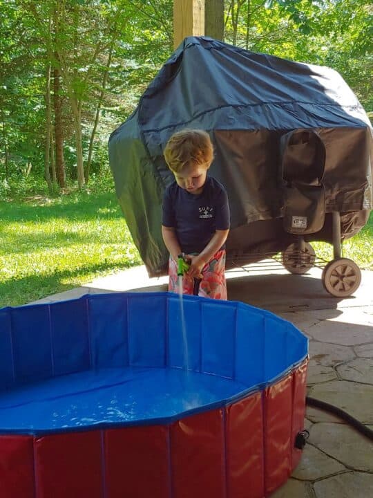 Toddler boy filling hard sided kiddie pool with hose
