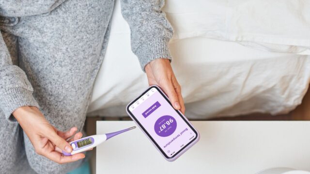 Woman holding basal body temperature thermometer syncing with fertility app.