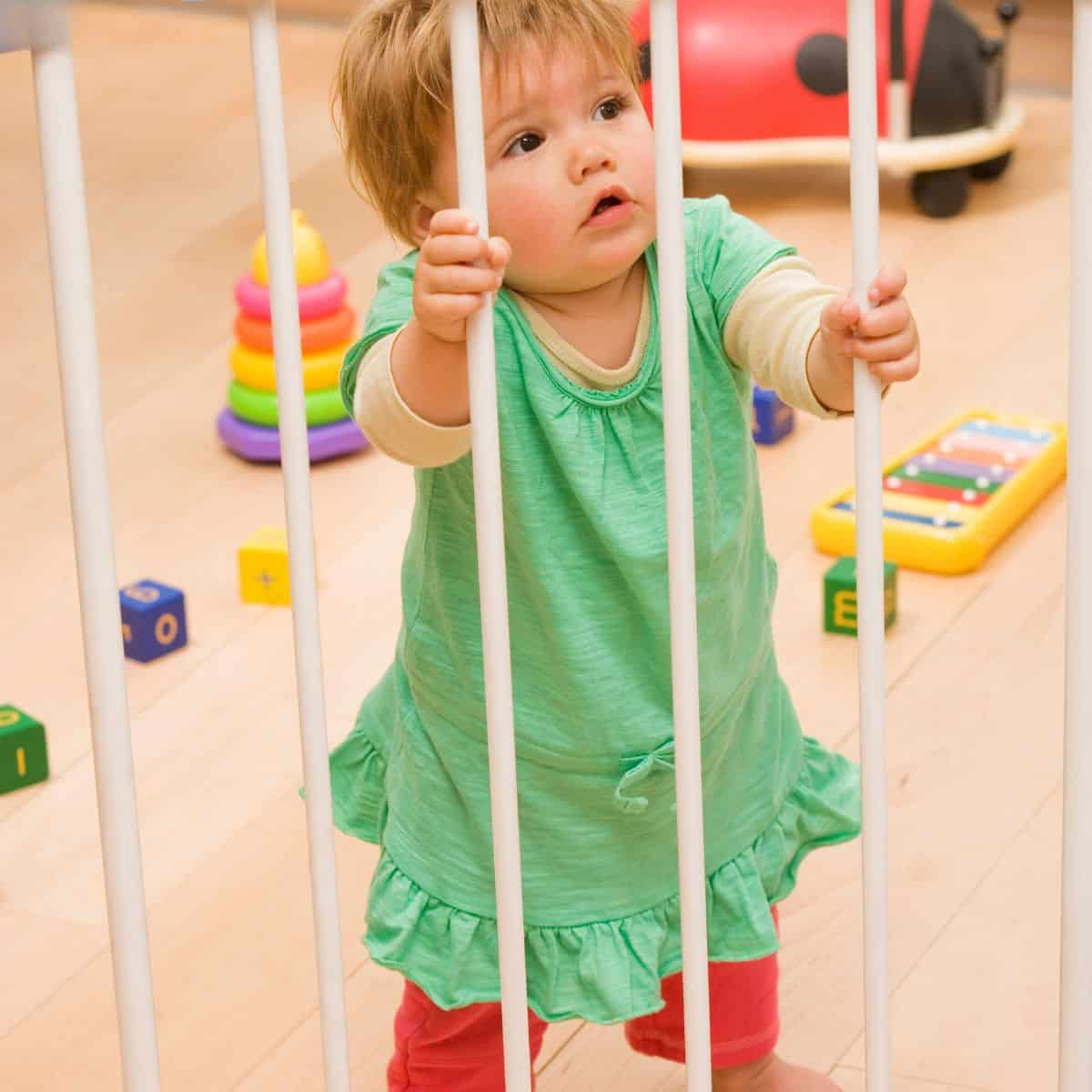 Baby standing at gate of playpen with toys in the background.